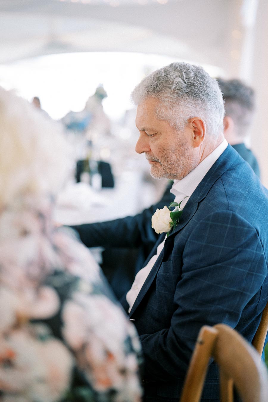 Elderly man in formal attire with a white rose boutonniere, seated at a wedding reception, engaged in conversation.