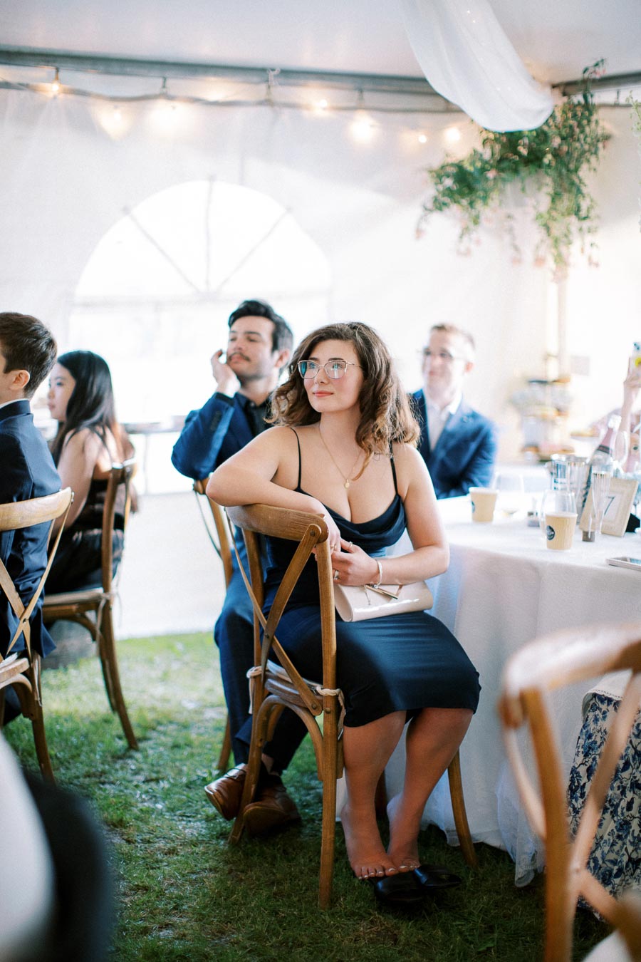 A group of elegantly dressed people sitting at a table in a wedding reception tent with festive lights and greenery. The focus is on a woman in a black dress attending the event, holding a purse and smiling attentively.