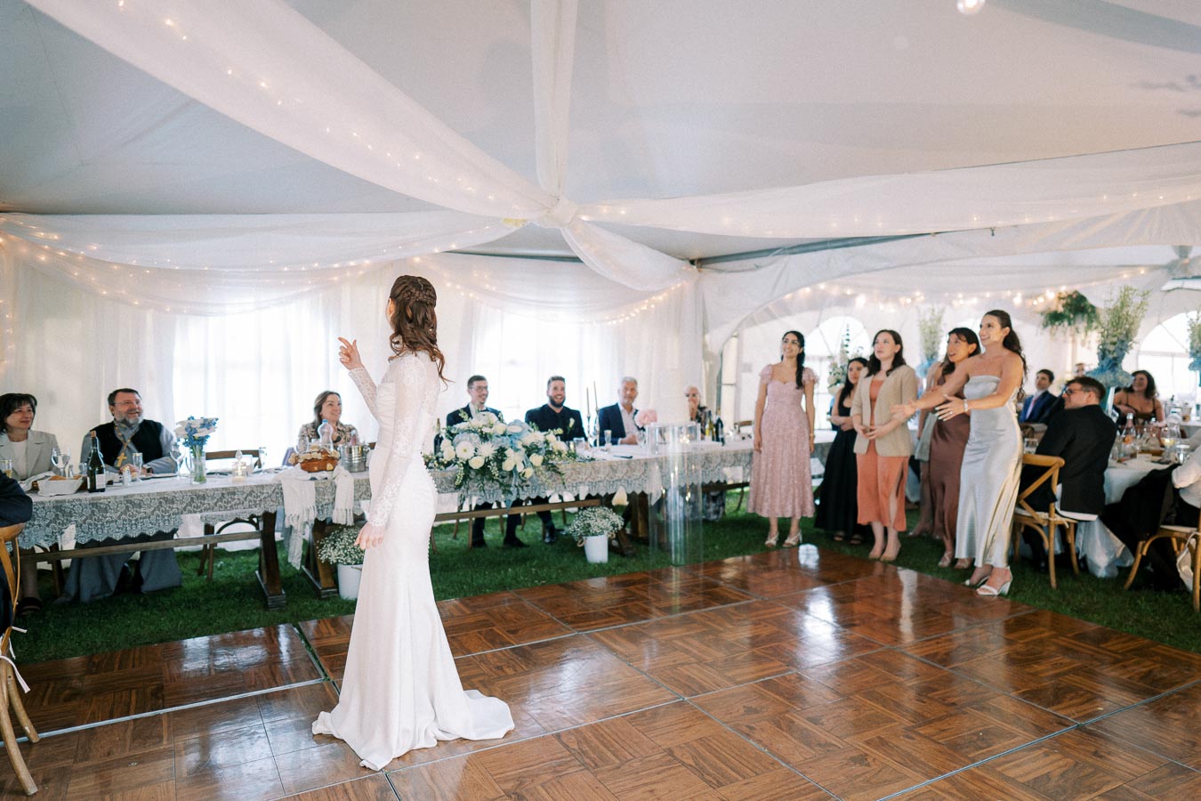 Bride in a white wedding dress stands on a dance floor inside a decorated wedding tent, gesturing towards a group of smiling guests in formal attire, with a table adorned with flowers in the background.