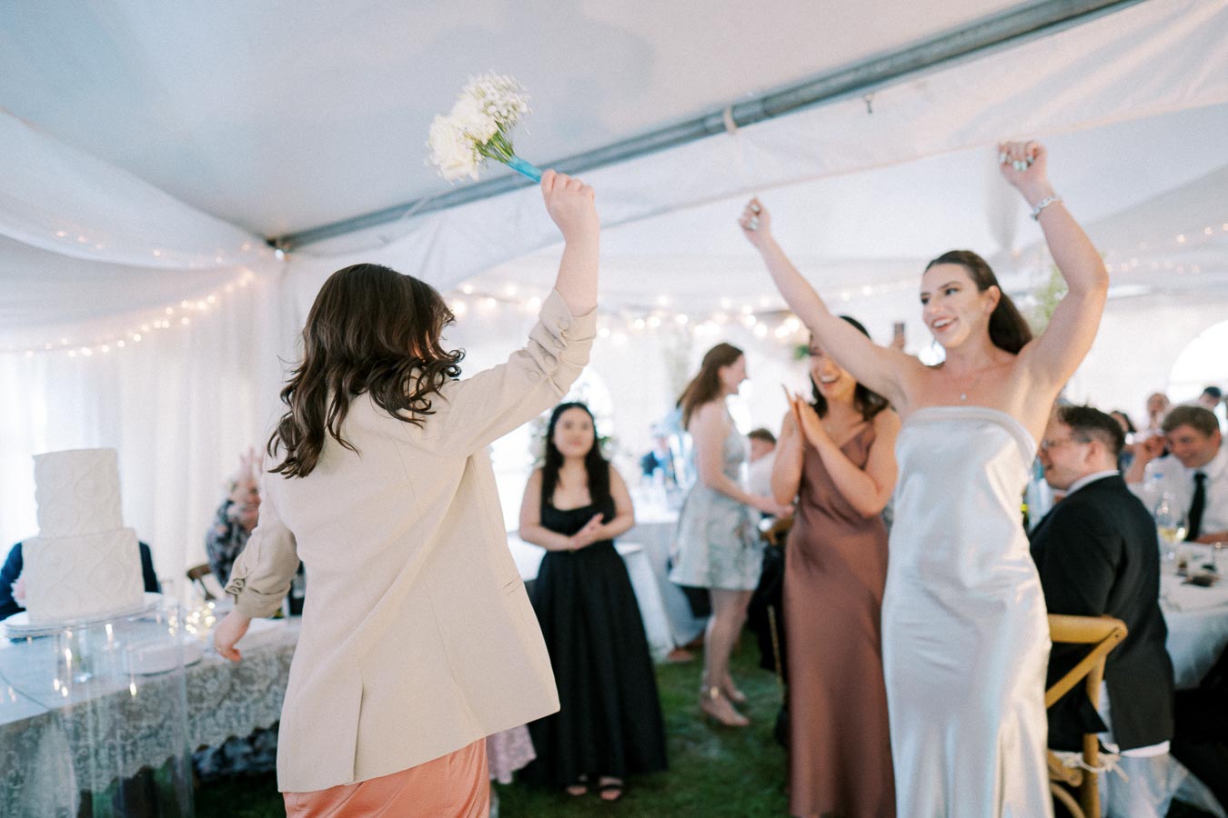 Wedding reception scene inside a tent, featuring a joyful moment where a woman in a satin dress raises her arms in celebration while another guest holds a bouquet, surrounded by attendees in formal attire.