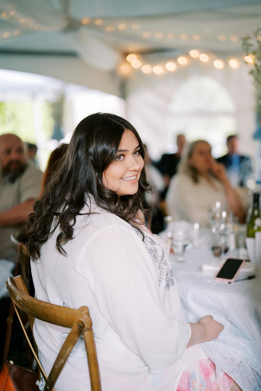 Smiling woman seated at a decorated indoor event with soft lighting and attendees visible in the background, capturing a joyful atmosphere.