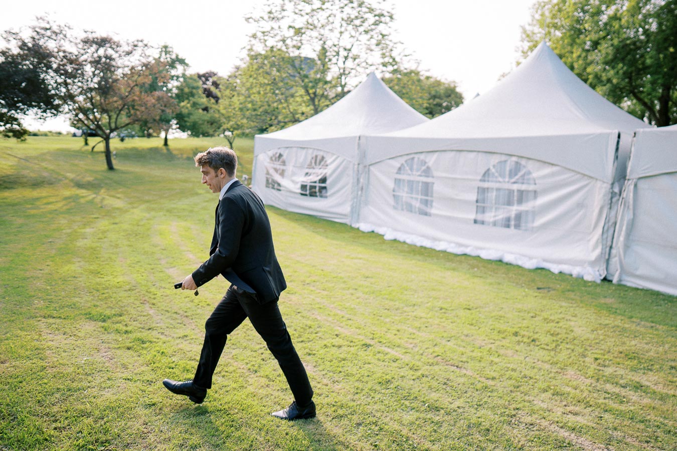 A man in a suit walking across a grassy field past large white event tents, with trees in the background on a sunny day.