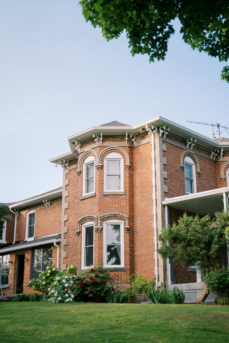 Historic red brick Victorian-style house with ornate detailing, surrounded by lush green lawn and colorful flower garden, under a clear blue sky.