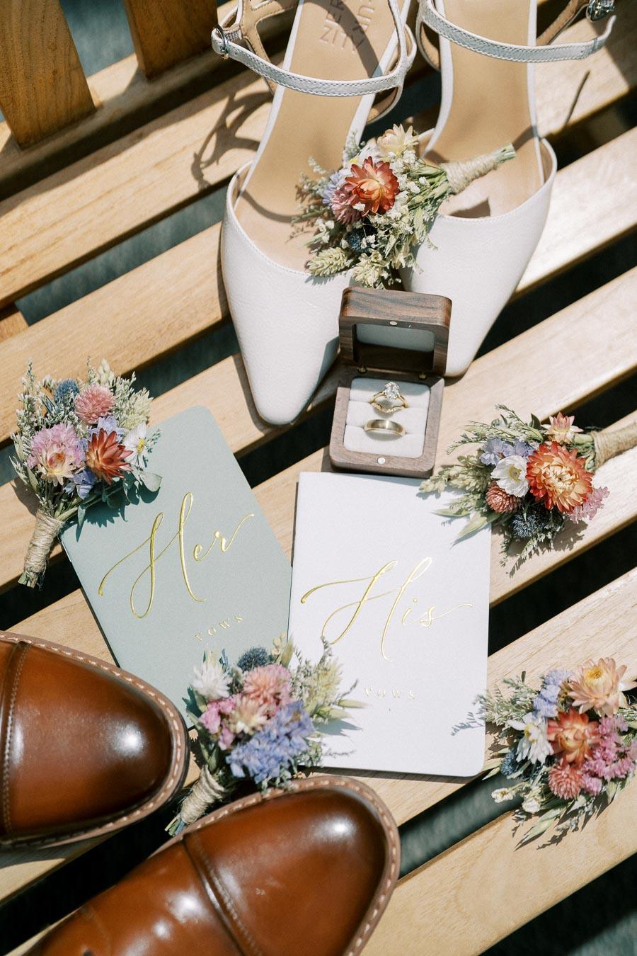 Elegant wedding flat lay featuring brown leather shoes, white bridal shoes with floral decorations, a pair of wedding rings in a wooden box, and His and Her vow booklets on a wooden bench.