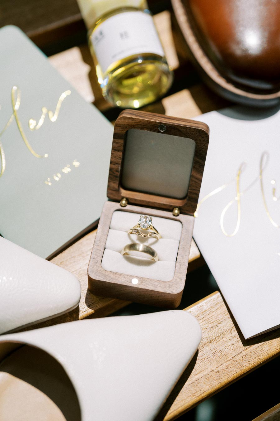 Elegant diamond engagement and wedding rings in an open wooden box, surrounded by bridal accessories including white shoes and 'His and Hers' vow booklets on a wooden table.