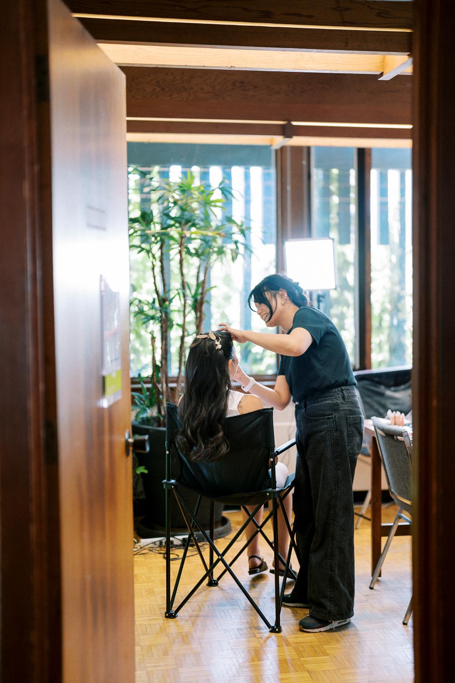 A hairstylist working on a client's hair in a well-lit room with large windows and indoor plants, creating a relaxed and professional atmosphere.
