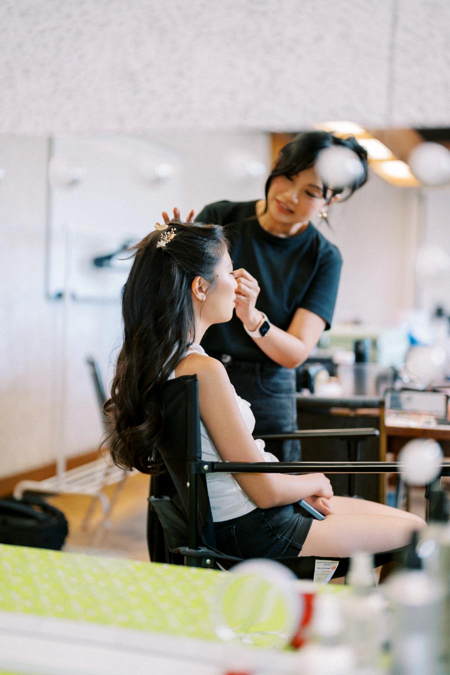 A makeup artist applying cosmetics to a young woman with long hair styled elegantly, seated in a salon chair. The setting is a professional beauty salon with a mirror reflecting part of the scene.
