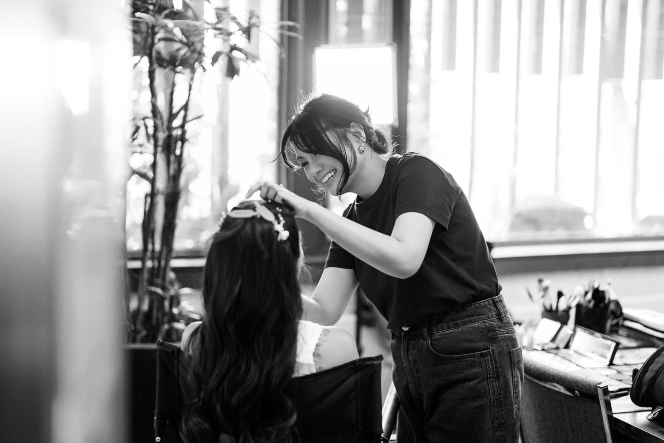 A hairstylist smiling while carefully styling a client's hair in a salon setting, with natural light coming through large windows.