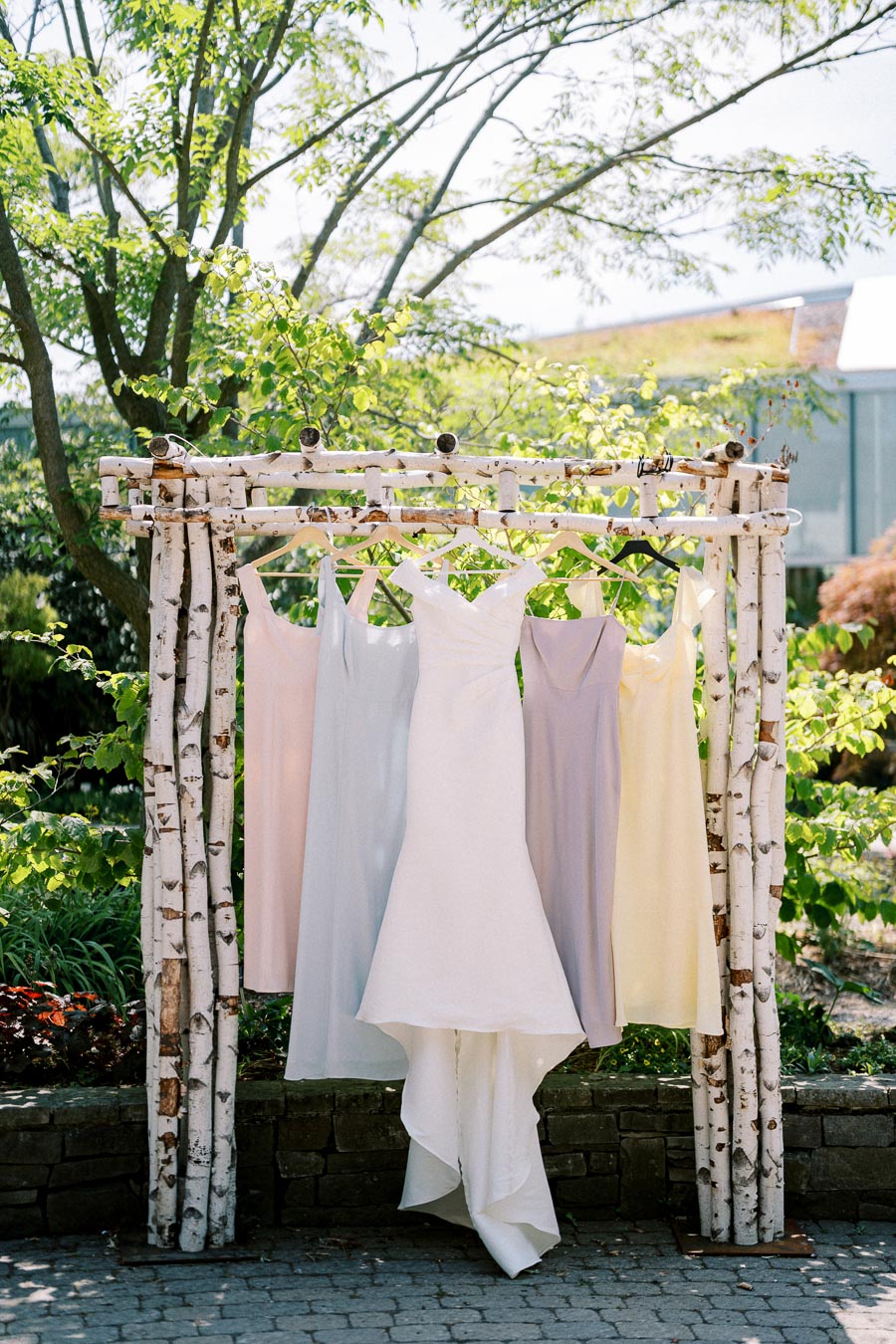 Elegant white bridal gown and pastel bridesmaid dresses hanging on rustic wooden frame, set outdoors against a lush green garden background.