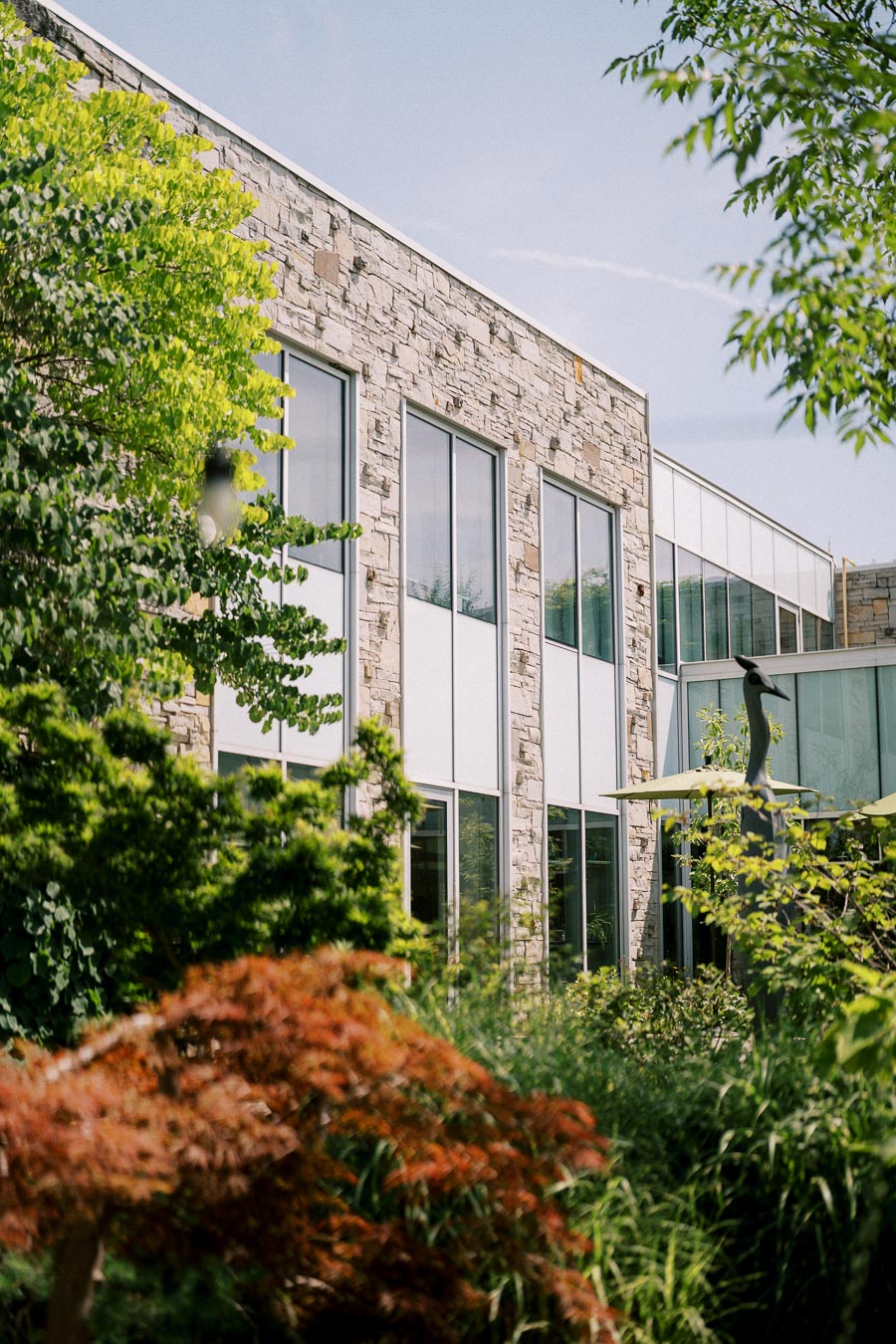 Modern building exterior with large glass windows and stone facade, surrounded by lush green trees and plants, under a clear blue sky.