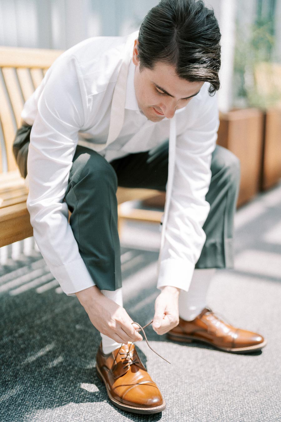 A man in a white shirt and dark trousers sitting on a bench ties the laces of his brown leather oxford shoes, with sunlight casting patterns on the floor.