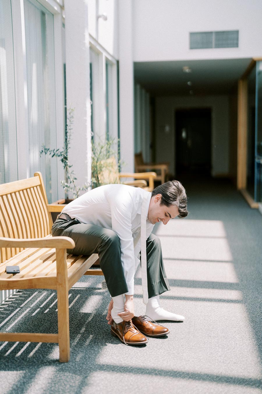 Man tying brown dress shoes while sitting on a wooden bench in a sunlit corridor, wearing a white shirt and dark pants, preparing for a formal event.