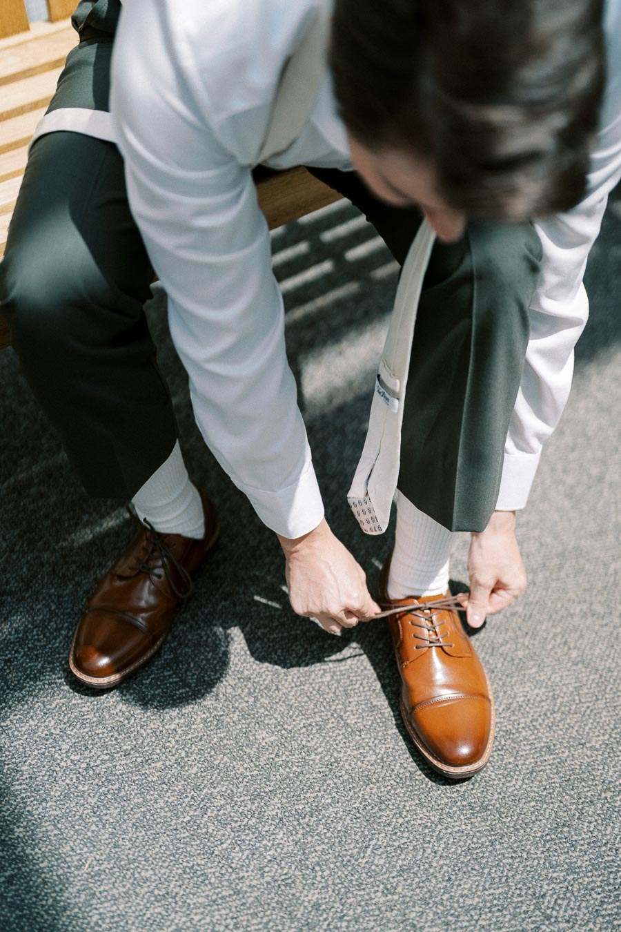 Person tying brown leather dress shoes on a bench, wearing a white shirt and dark trousers.