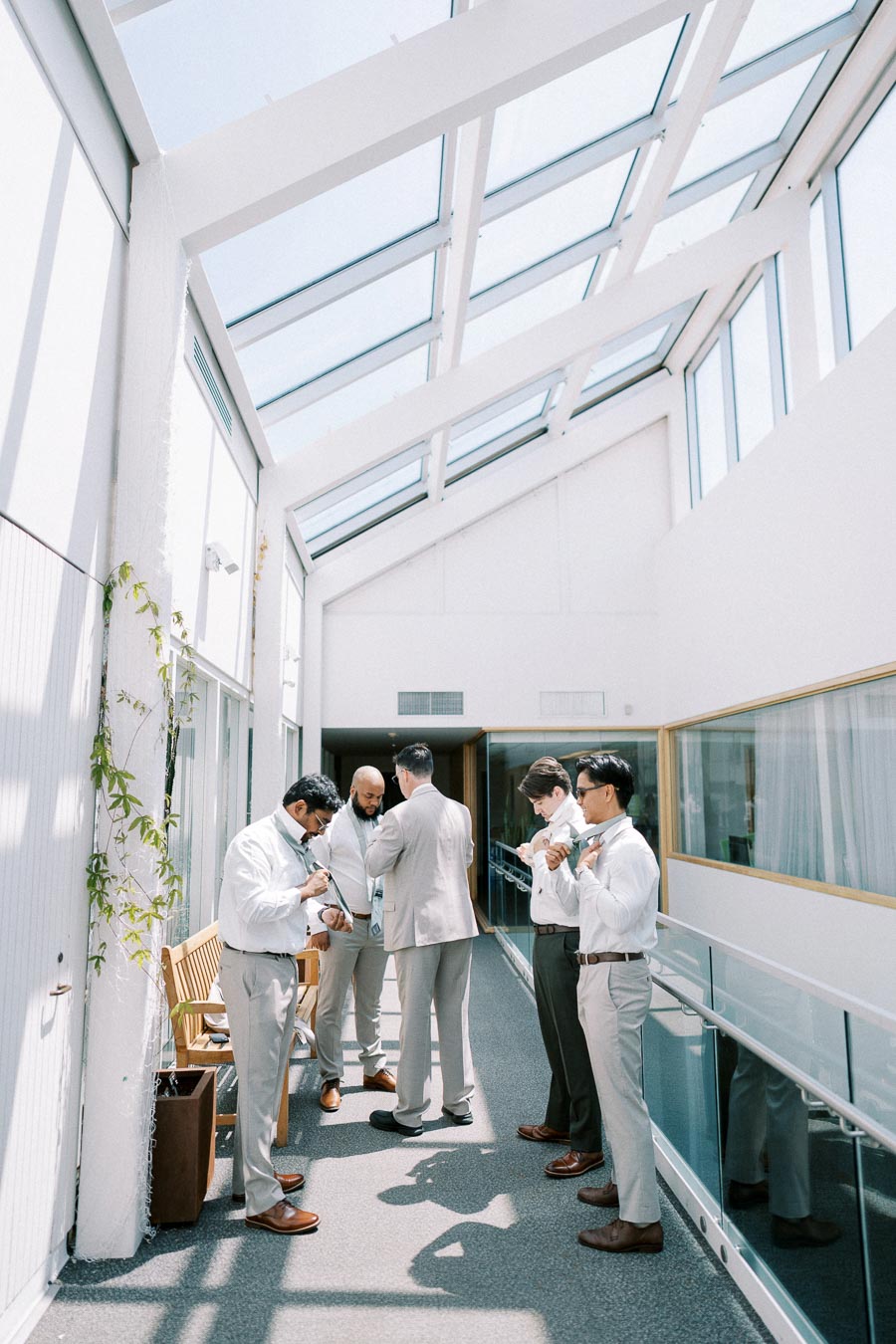 A group of men preparing for a formal event in a bright, sunlit hallway with a glass ceiling, wearing light-colored suits and adjusting their ties.