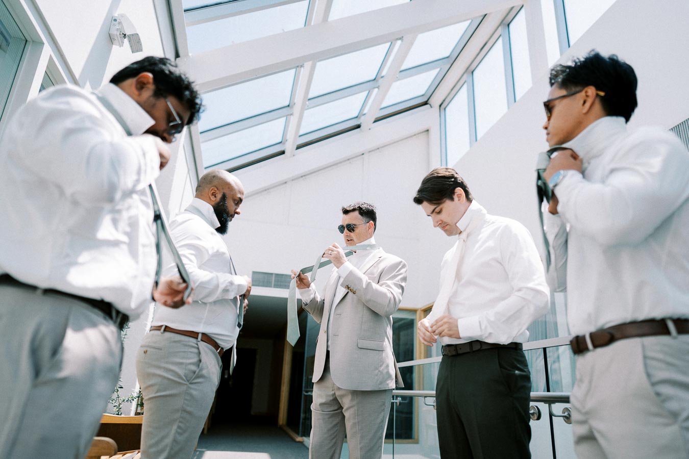 Group of men in formal attire adjusting ties in a well-lit room with large windows.