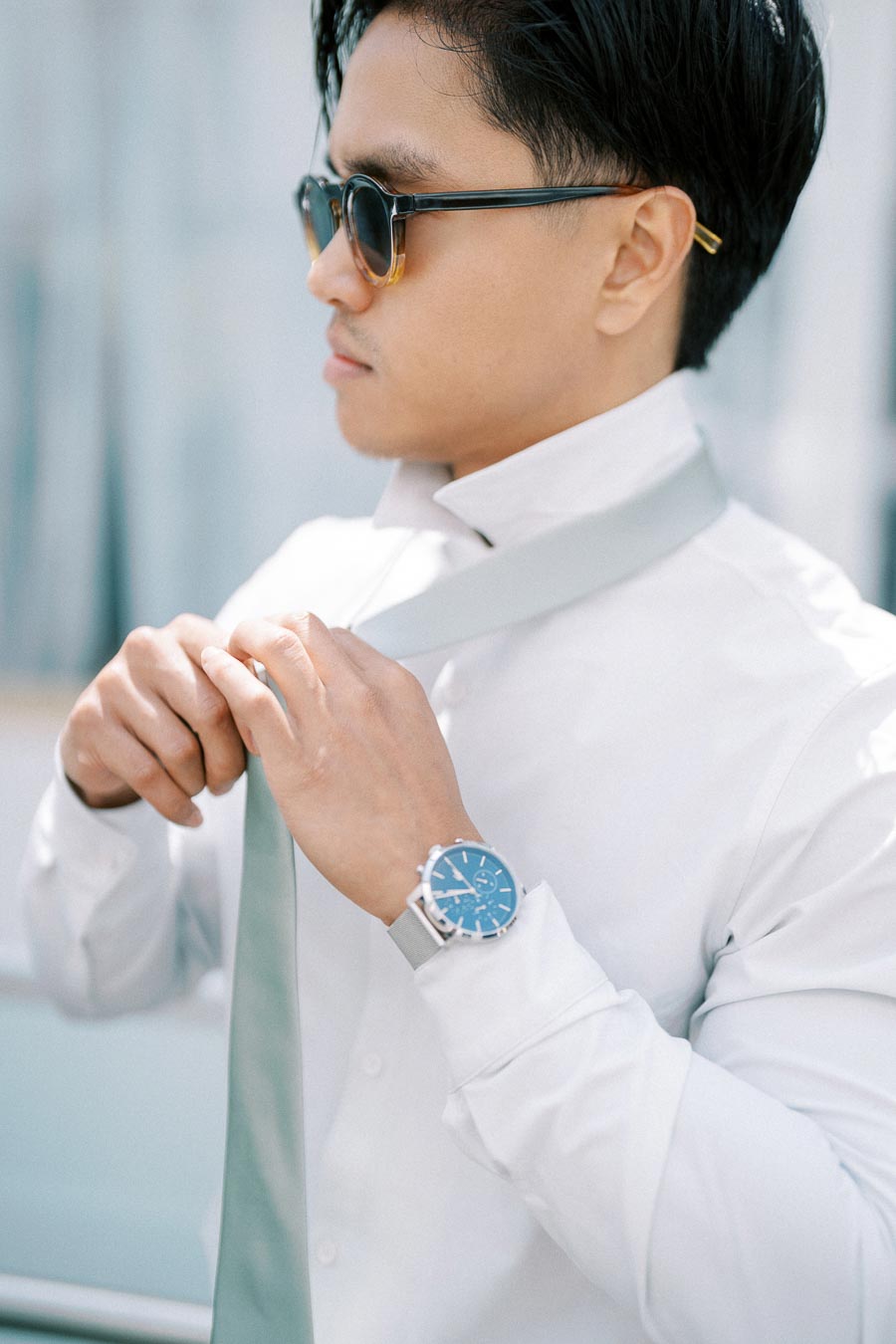 Man in sunglasses and a white shirt adjusting a light gray tie, wearing a stylish watch with a blue face, outdoor setting.