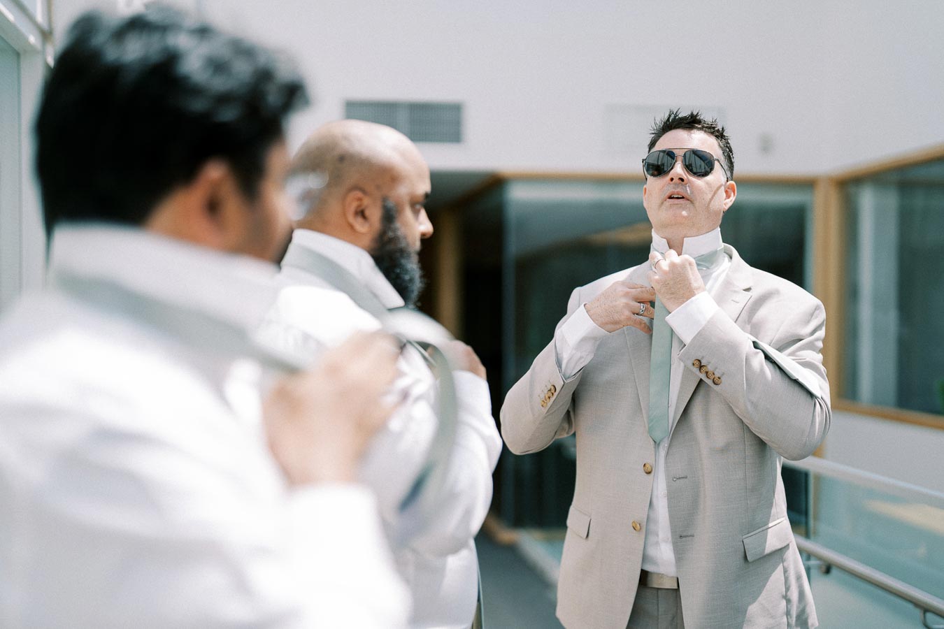 Three men in suits adjusting their ties, with one wearing sunglasses, preparing for a formal event indoors.