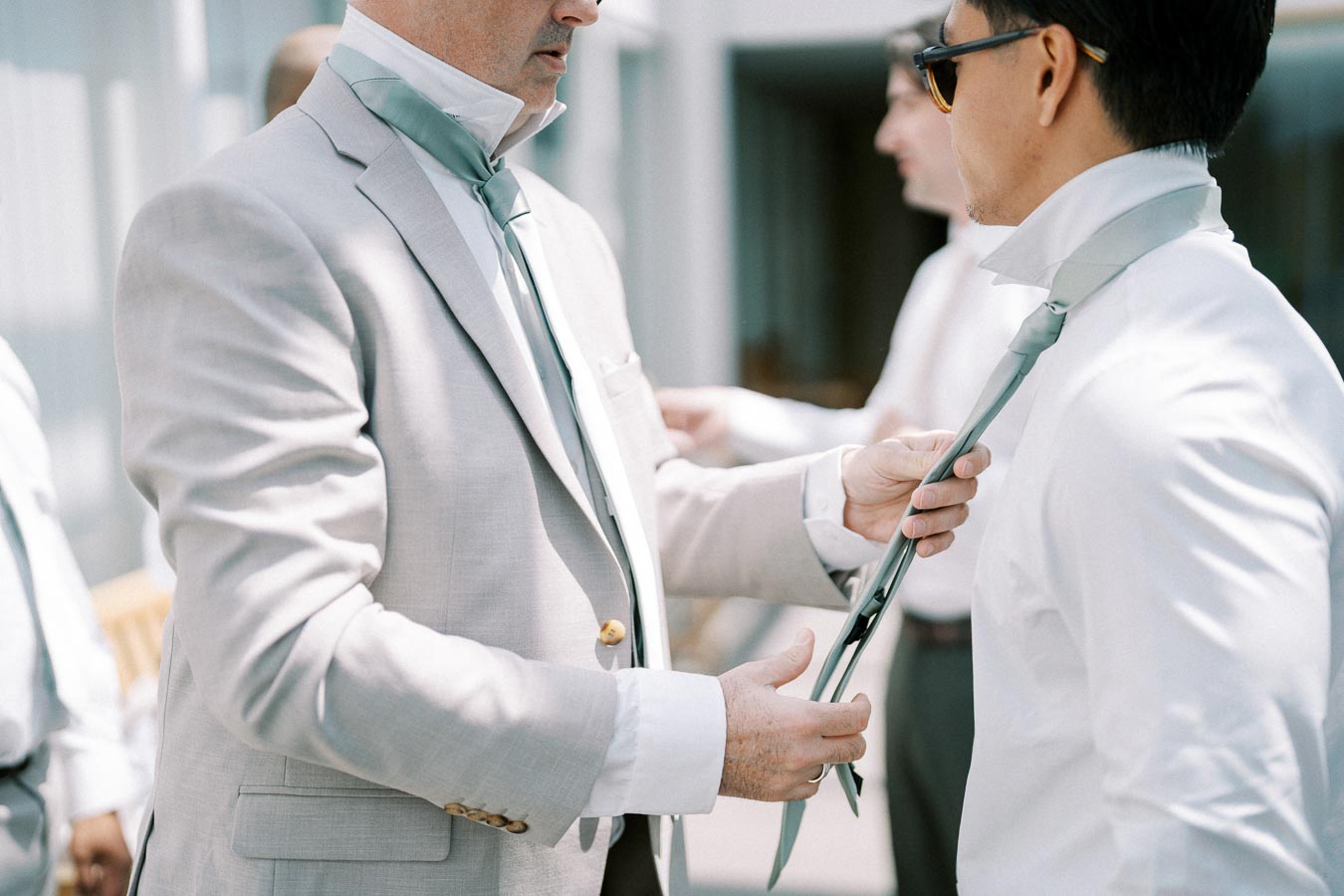 Two men in light-colored suits adjusting neckties in preparation for a formal event.