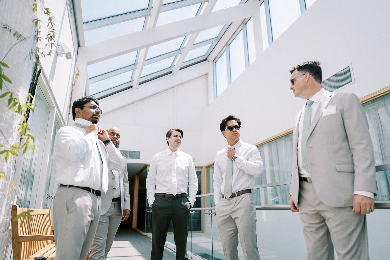 Group of five men in light-colored suits standing together in a sunlit glass atrium, adjusting ties and conversing.