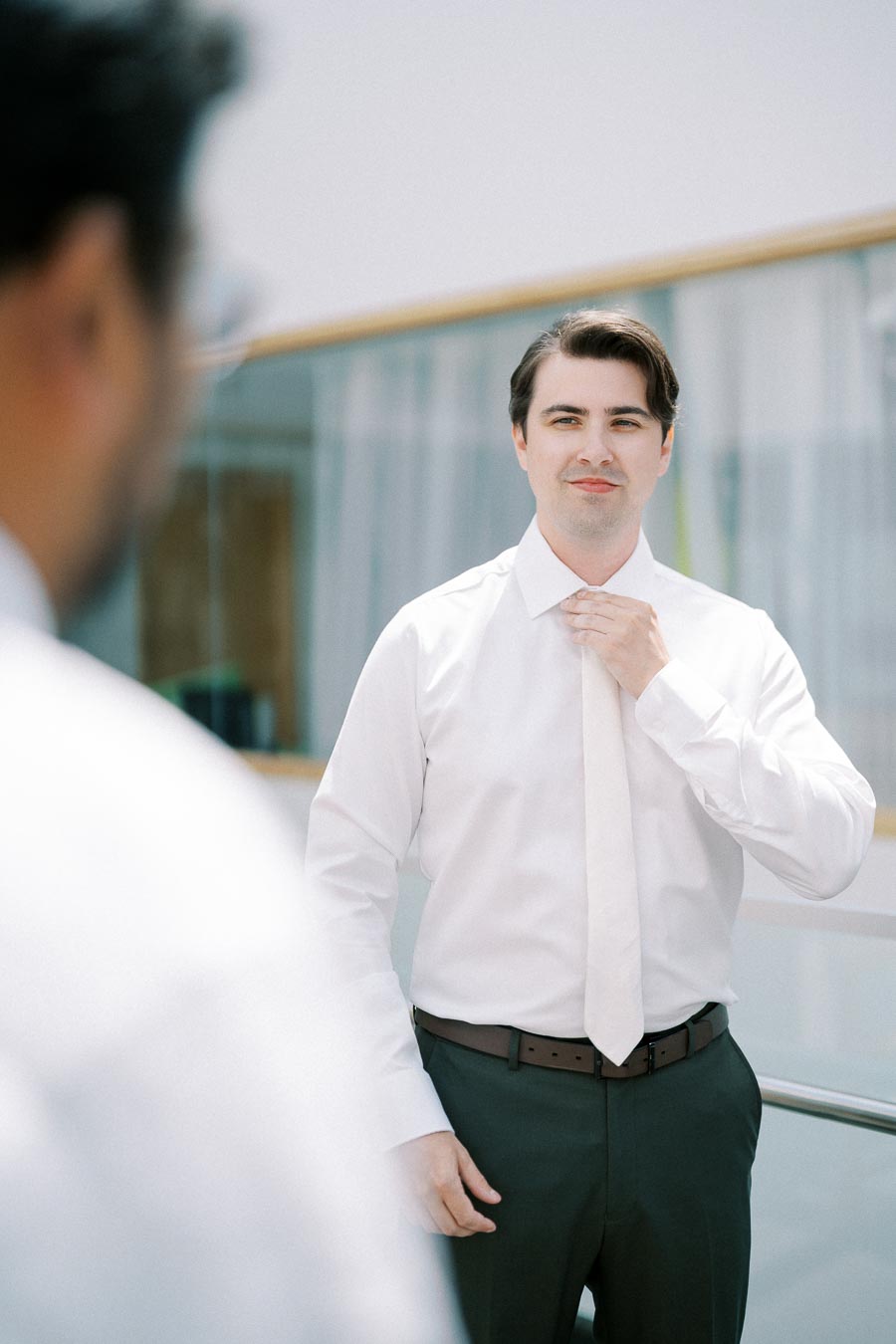 A groom adjusting his tie in a bright room, preparing for a wedding ceremony.