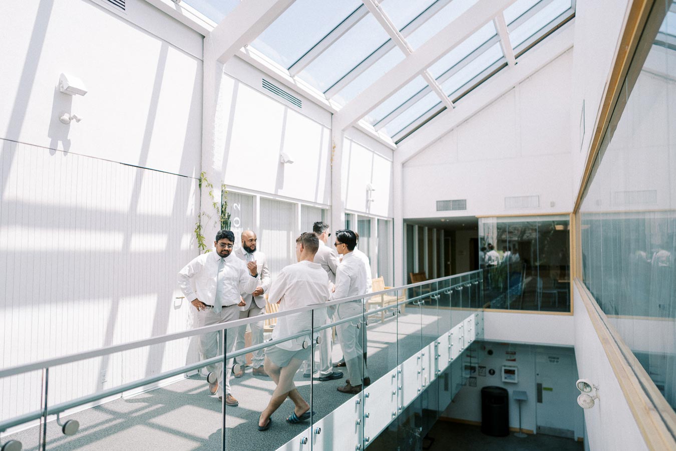 Group of men in formal attire having a conversation in a brightly lit modern hallway with glass windows and railings.
