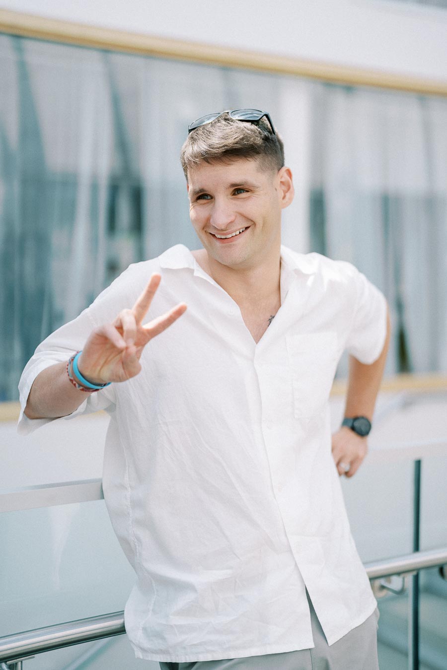 Smiling person in a white shirt making a peace sign while leaning on a railing outdoors.