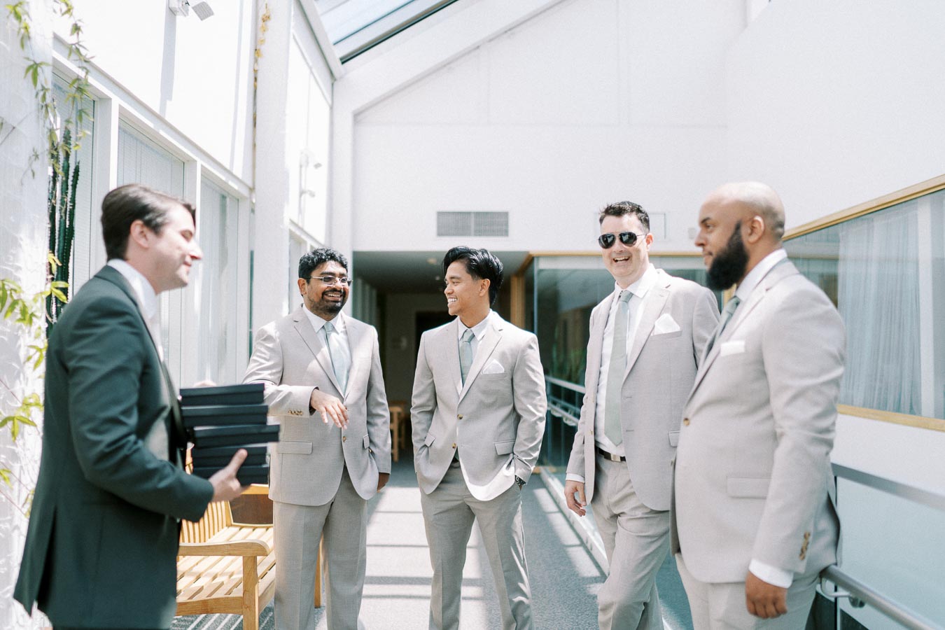 Group of men in light gray suits standing and conversing in a bright, modern hallway, with one holding a stack of black boxes, captured in a candid moment.