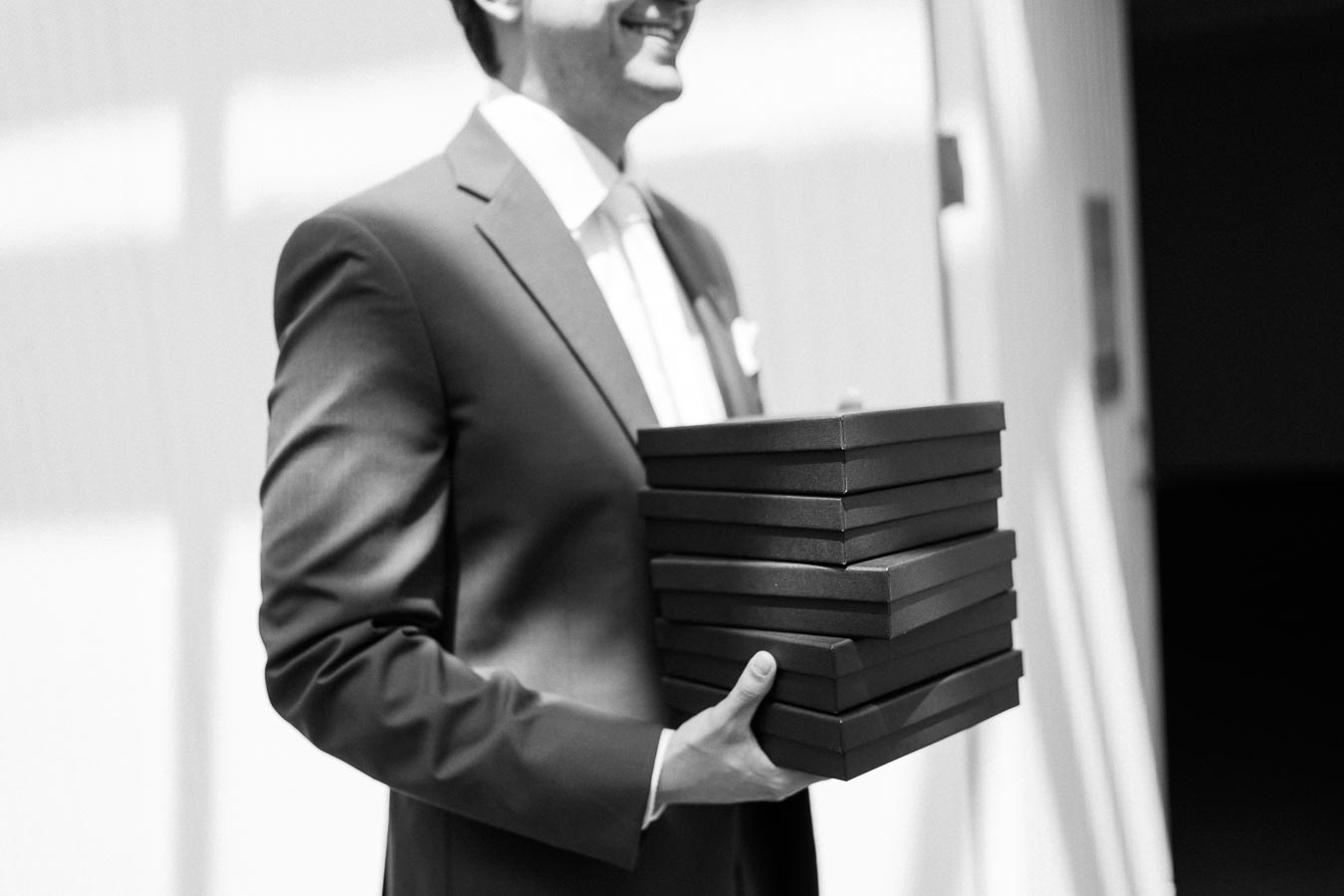 Black and white image of a person in a suit smiling while holding a stack of gift boxes, highlighting a professional and celebratory occasion.