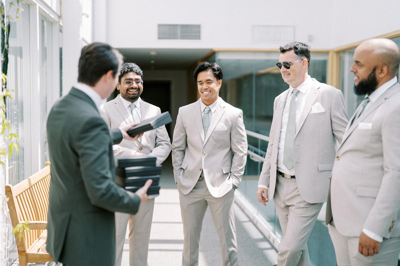 A group of groomsmen in light gray suits and ties smiling and standing in a sunlit hallway, preparing for a wedding ceremony.