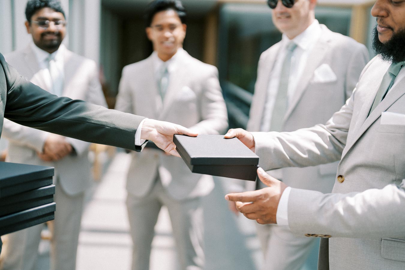 Groomsmen in light gray suits exchanging gift boxes during a wedding ceremony.