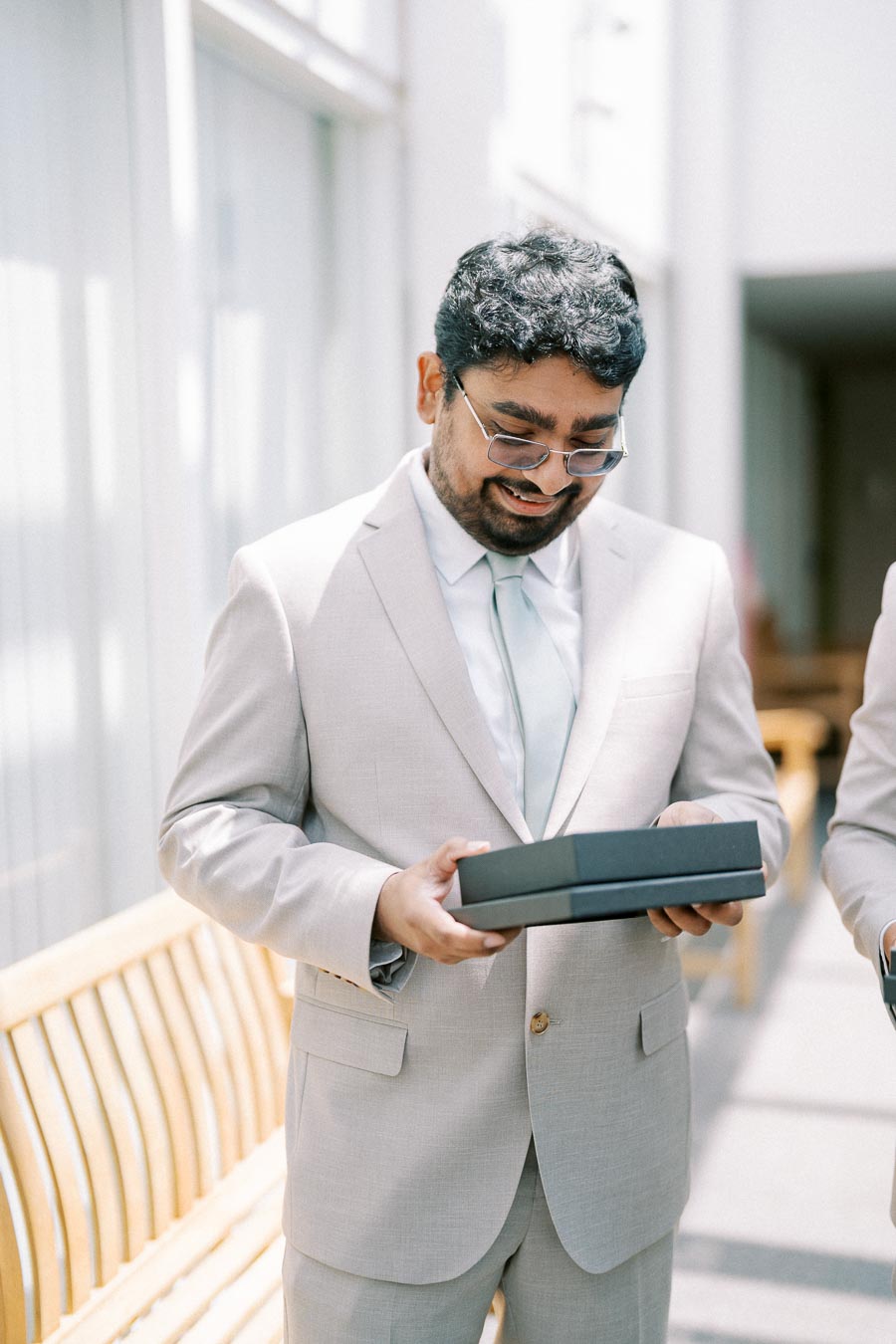 Man in light-colored suit smiling while holding a gift box indoors.