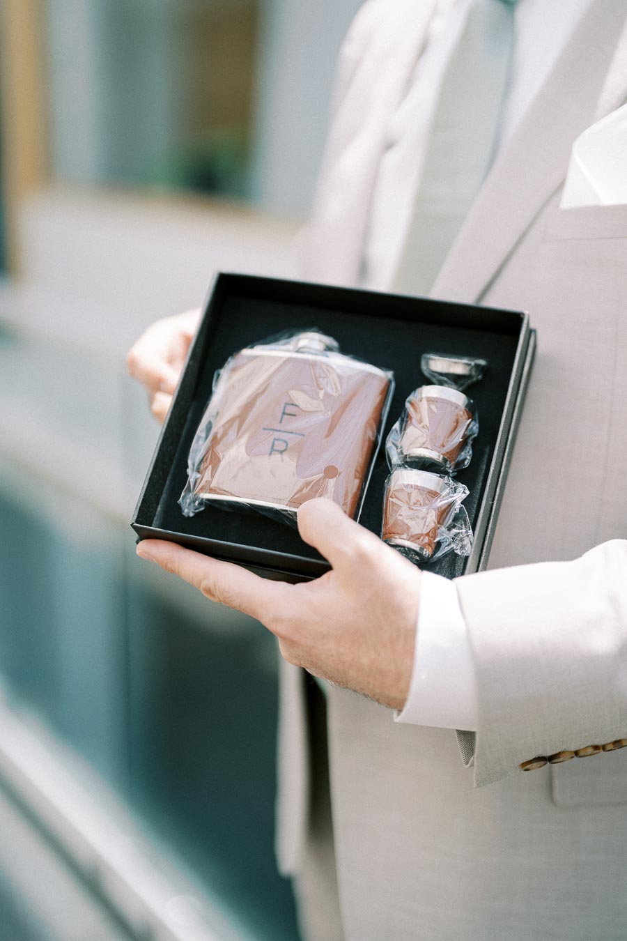 A person in a light suit holding a gift box containing a personalized flask and shot glasses, wrapped in plastic, ideal for a groomsman or special occasion gift.