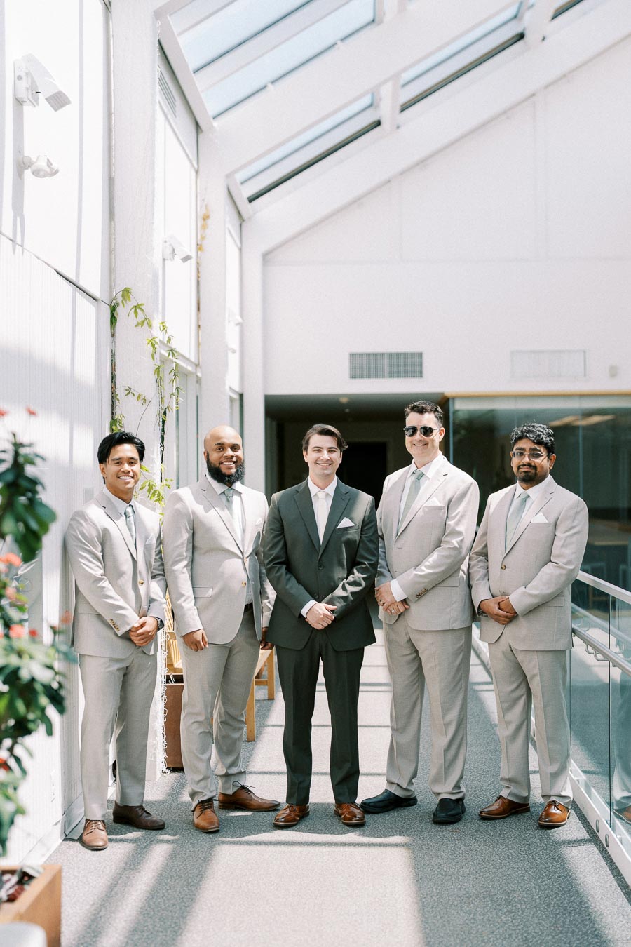 Groom and groomsmen in light suits posing indoors near a sunlit glass hallway.