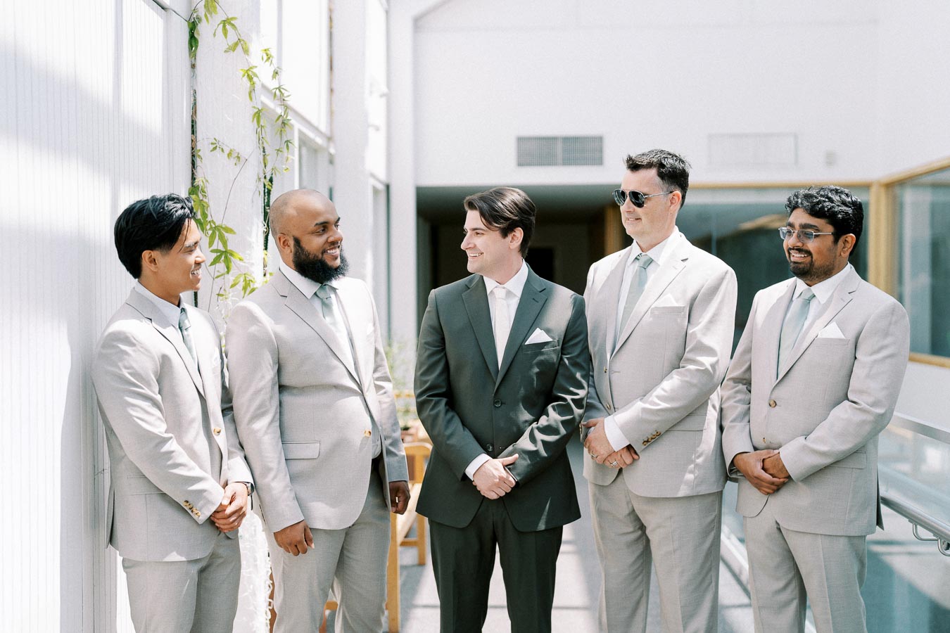 Groom and groomsmen in light grey suits and ties smiling together in a bright, modern venue.