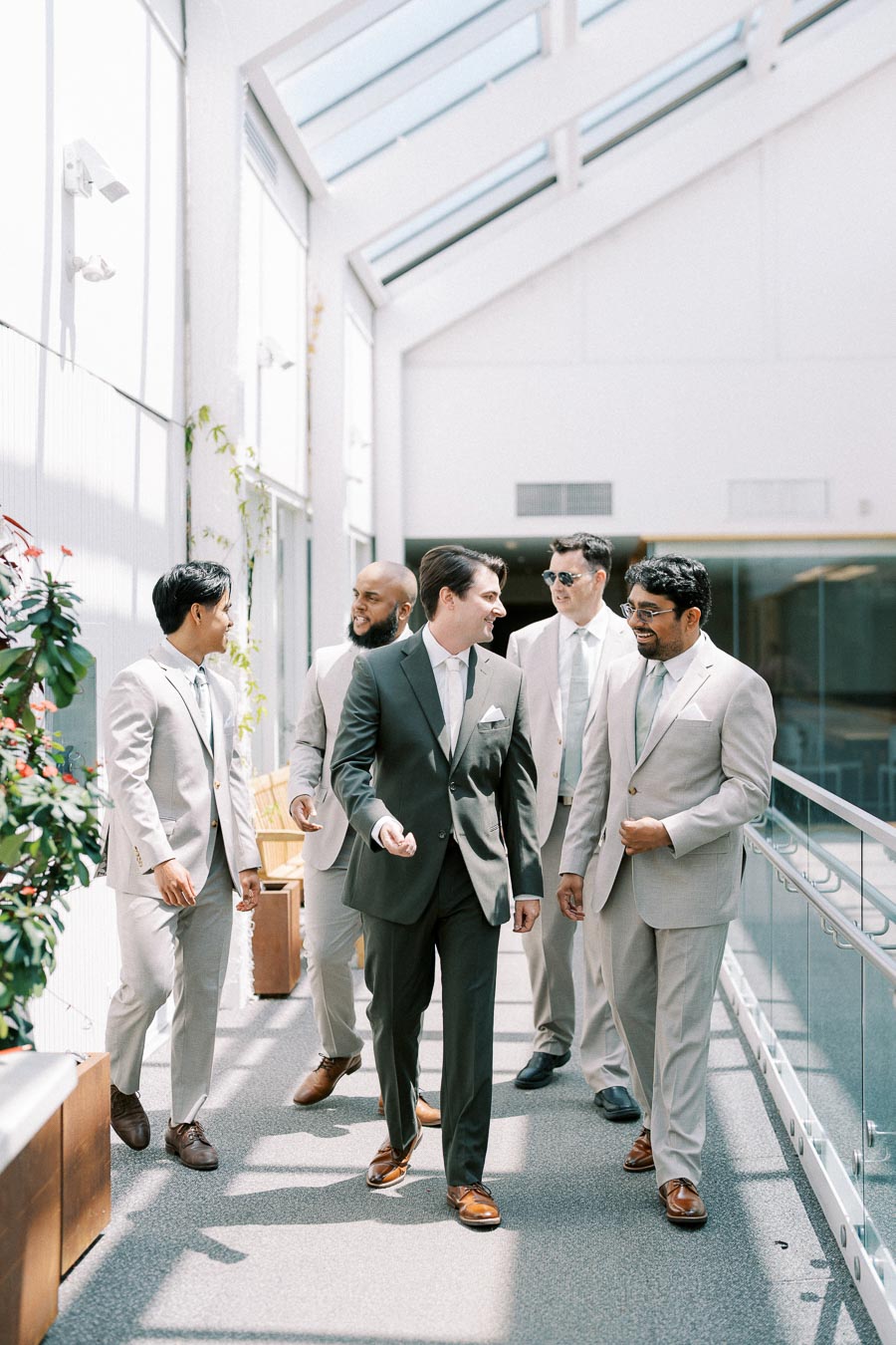 Five men in suits walking and conversing in a sunlit corridor, exuding a confident and professional atmosphere, perfect for a modern wedding or business event.