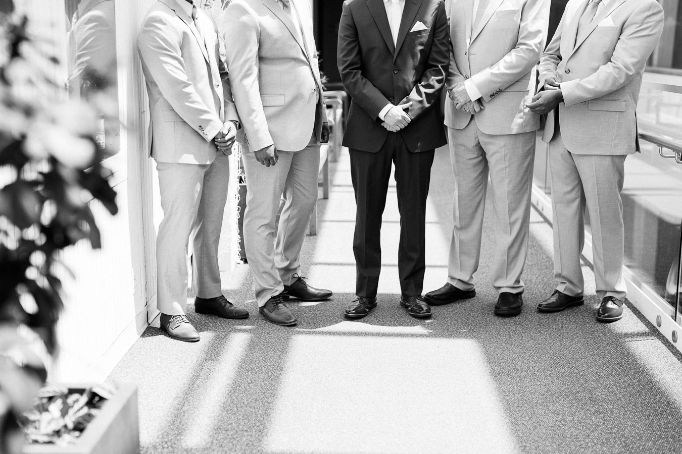 Black and white image of five men in suits, standing indoors, with light streaming through windows, captured in a stylish composition focusing on lower bodies and legs.