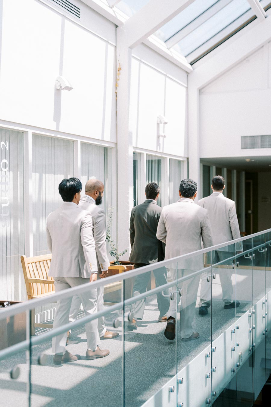 Group of five men in formal suits walking down a sunlit corridor with glass railings and white walls.