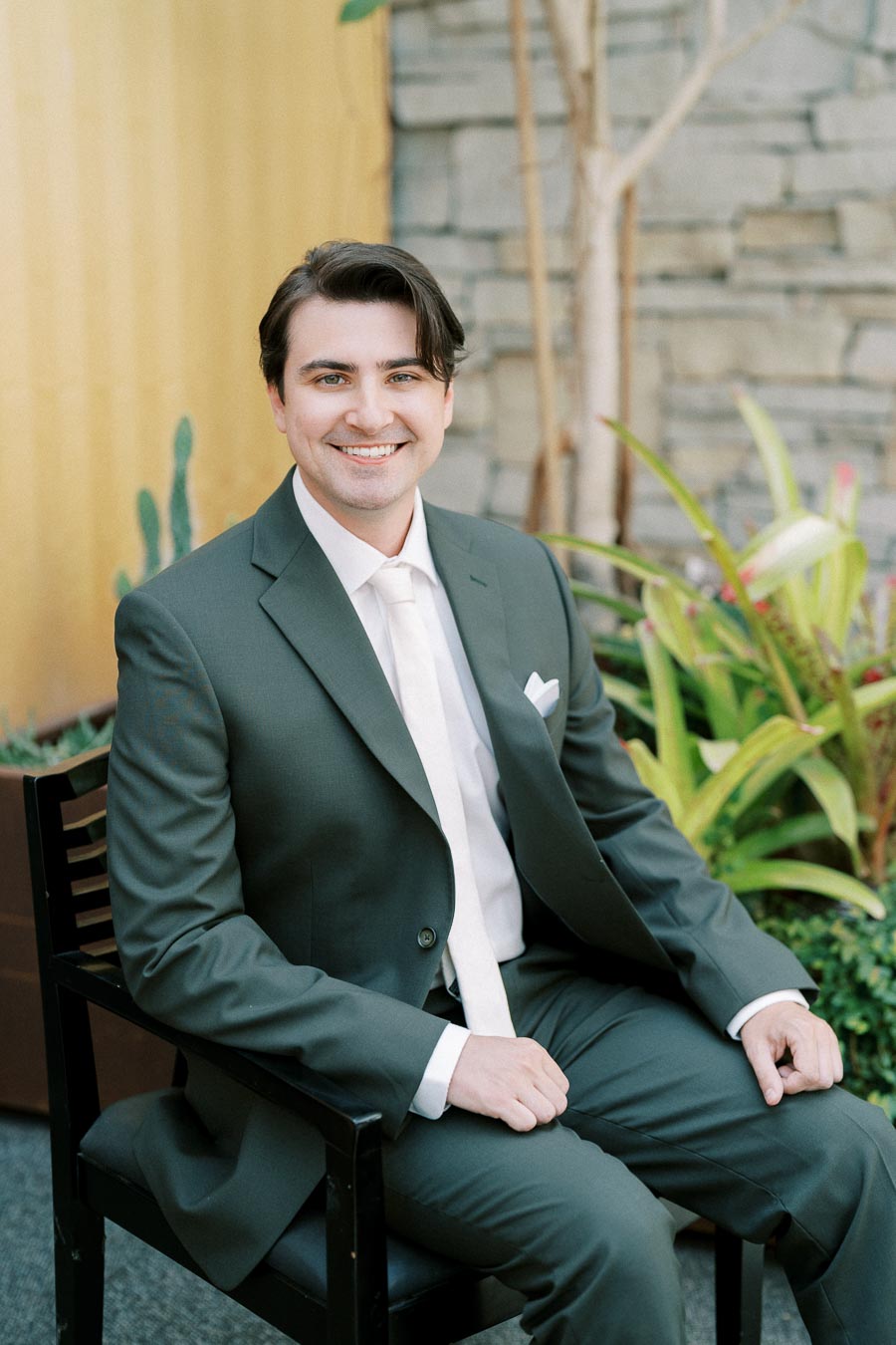 Man in a green suit sitting on a chair in a garden setting with plants and stone wall background, smiling at the camera.