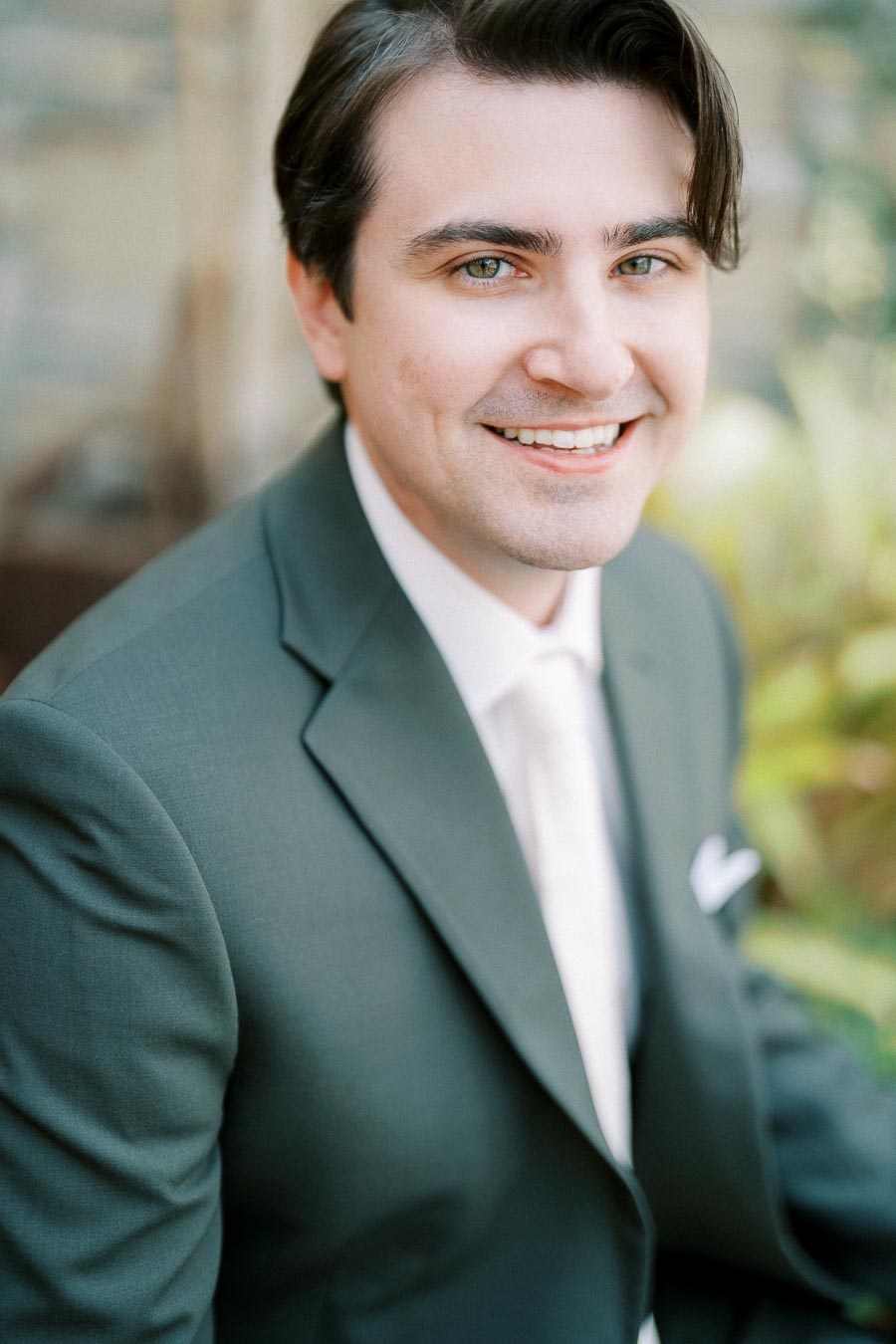 Smiling man in a dark green suit with a white shirt, sitting outdoors with a blurred natural background. ```