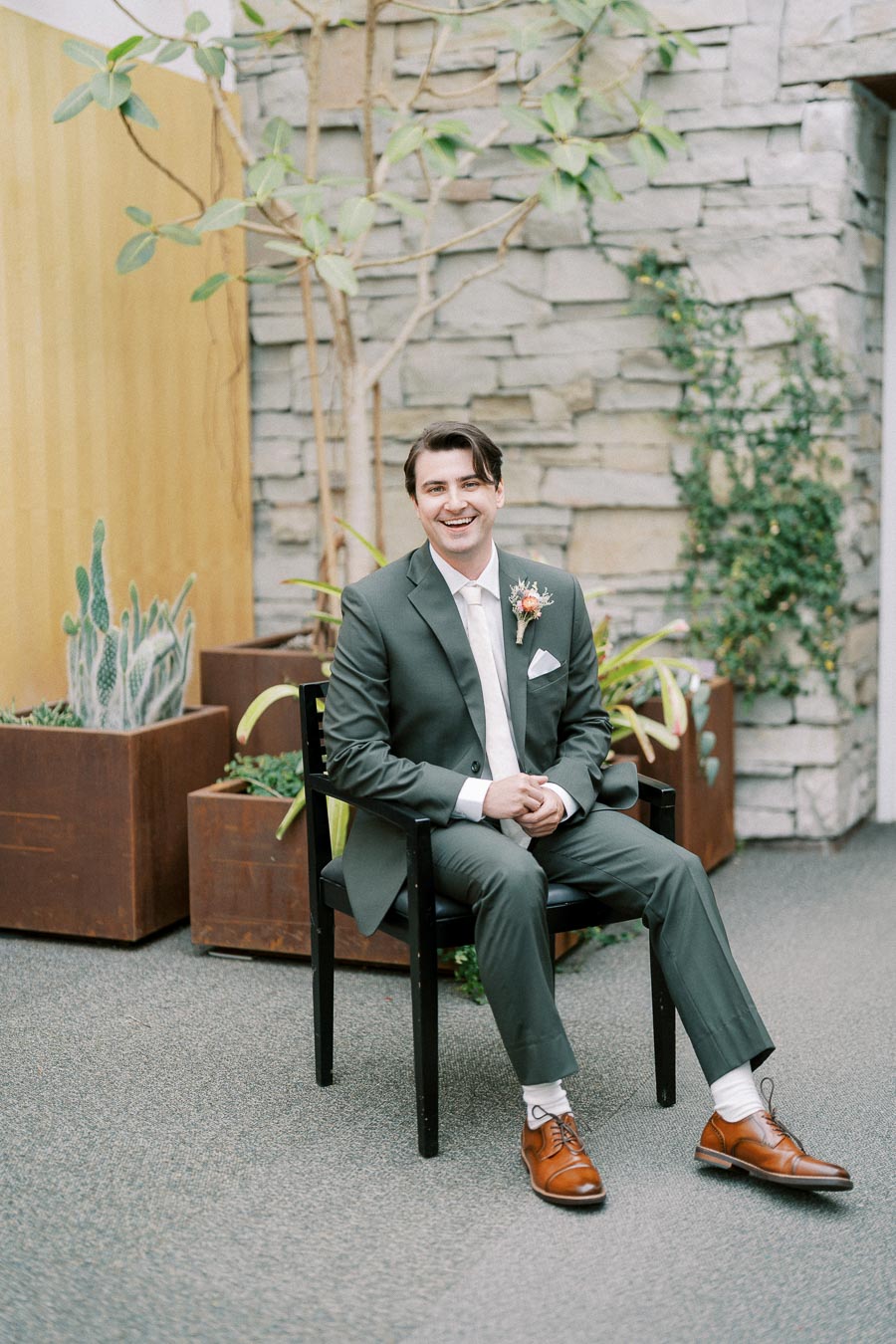 A smiling groom in a green suit sits on a black chair in an indoor setting with stone and plant decor, featuring brown dress shoes.