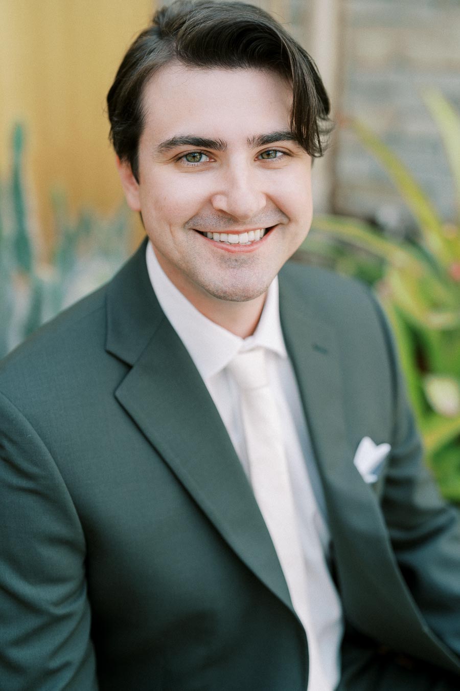 A smiling man in a dark suit and white shirt, seated outdoors with a blurred natural background, exuding a professional and approachable demeanor.