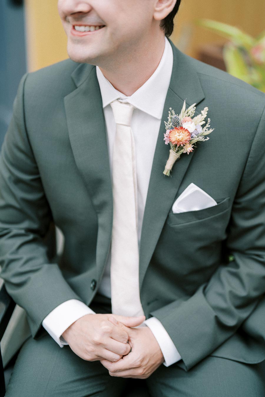 A groom in a tailored green suit with a floral boutonniere sits smiling, highlighting elegant wedding attire with attention to detail.
