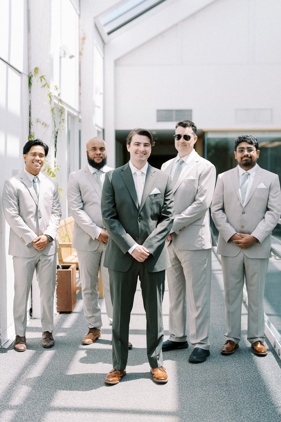 A group of five groomsmen in light gray suits and brown shoes standing together indoors, smiling warmly, with natural light streaming in through large windows.