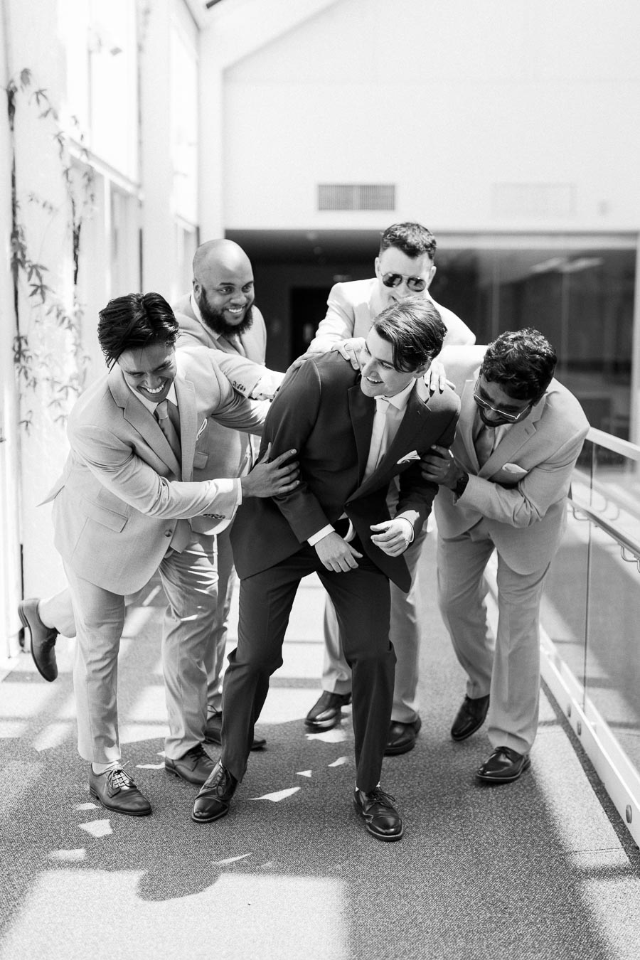 A group of five groomsmen in suits laughing and playfully interacting with each other in a sunlit hallway, captured in black and white.