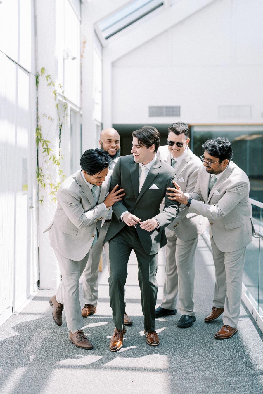 Groom in dark suit smiling and laughing with groomsmen in light gray suits, walking in a bright hallway, celebrating before the wedding ceremony.