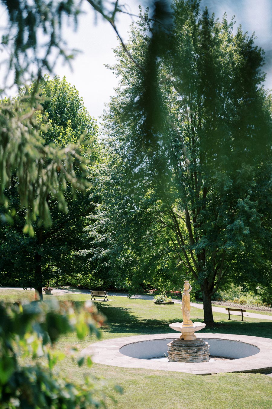 Tranquil park scene featuring a stone fountain with a statuesque figure in the foreground, surrounded by lush green trees and benches under a clear blue sky.