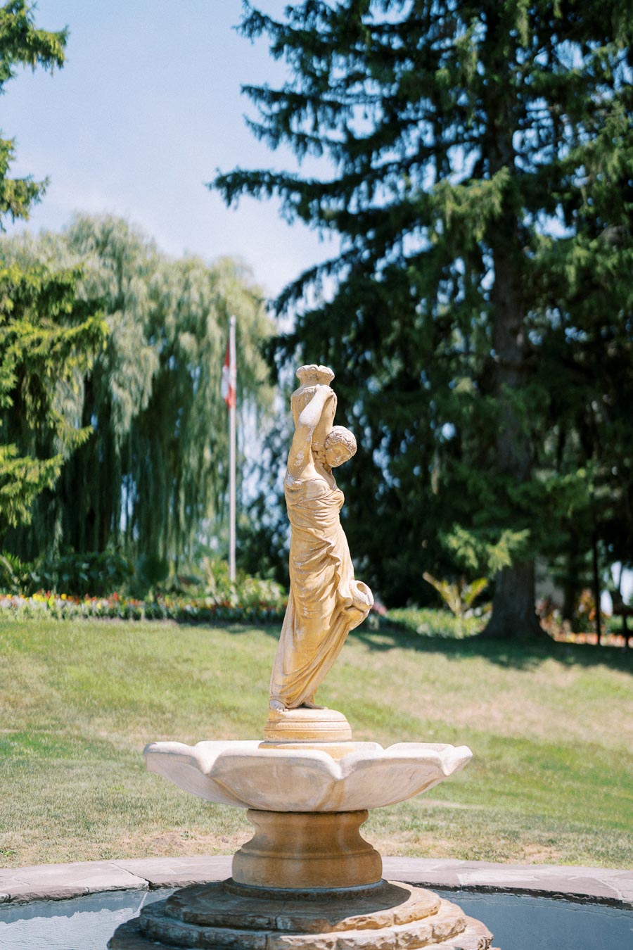 Elegant stone statue fountain in a lush park setting, surrounded by greenery and trees, under a clear blue sky.