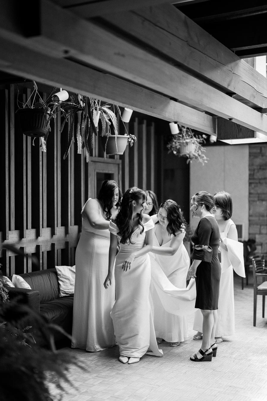 Black and white photo of a bride surrounded by bridesmaids. They're helping her adjust her gown in a beautifully decorated indoor space with hanging plants. Ideal for content about weddings, bridal preparations, or wedding photography.
