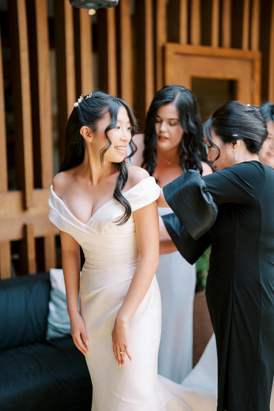 Bride in an elegant white off-the-shoulder wedding dress, smiling as bridesmaids assist her in preparation for the ceremony, set in a beautifully lit indoor venue.