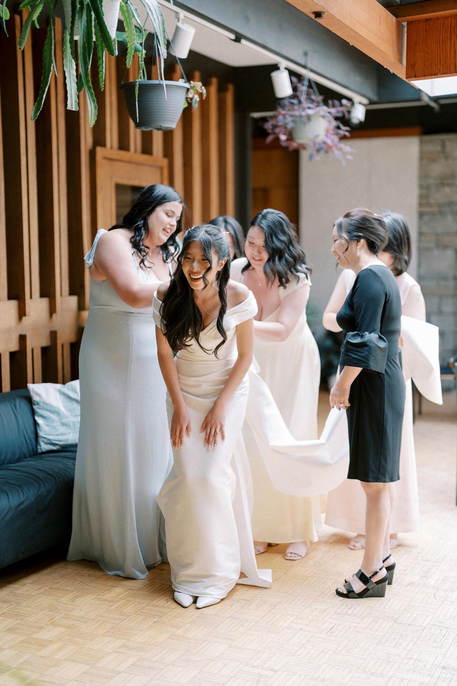 A group of bridesmaids assisting a joyful bride in a white wedding dress, preparing for her big day, in a modern venue with elegant decor and wooden accents.